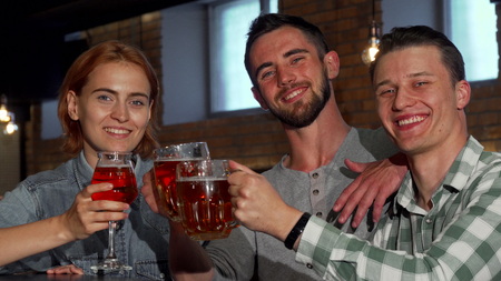 Happy young people clinking their beers and smiling to the camera. Group of friends enjoying drinking beer together at the pub. Cheerful woman and her male friends partying. Leisure concept.の写真素材