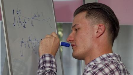 Handsome young man solving math problem on whiteboard. Attractive male student smiling to the camera while solving algebra equation on whiteboard at classroom. Education conceptの写真素材