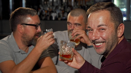 Happy mature man smiling to the camera while drinking whiskey with friends. Group of male friends having fun at the bar, drinking alcohol. Selective focus on a man smiling holding up his glass.の写真素材
