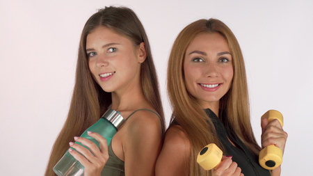 Two fitness women smiling at each other, posing with water bottle and dumbbells. Beautiful red haired sportswoman holding weights, smiling at her friend, standing back to backの写真素材