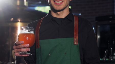 Unrecognizable brewer in apron showing thumbs up, holding glass of beer. Cropped shot of a happy...