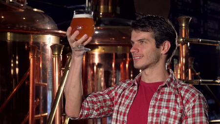 Cheerful male brewer smiling to the camera, examining beer in a glass. Brewery worker standing in...