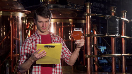 Young man with clipboard working at craft brewery, examining beer. Attractive cheerful male brewer enjoying working at his small brewery. Entrepreneurship, craft beer conceptの写真素材