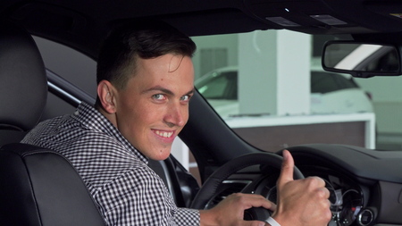 Handsome man smiling over his shoulder showing thumbs up, sitting in a new car. Male customer...