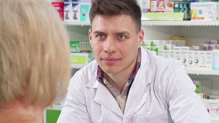 Male chemist listening to his senior female customer request on medicine. Sliding shot of a handsome pharmacist working at the drugstore, helping elderly clientの写真素材