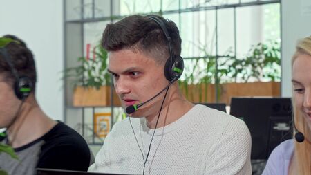 Young man working at customer support call center, wearing headset. Handsome male call center worker typing on computer. Friendly male operator helping customers onlineの写真素材