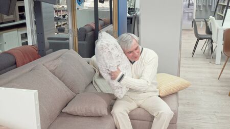Cheerful elderly man trying soft cushions on sale at furniture store. Happy senior man sitting on a couch at furnishings store. Elderly male customer at the furniture storeの写真素材