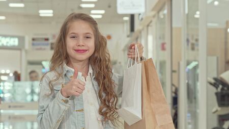 Happy cute little girl showing thumbs up at the shopping mall. Charming long haired girl holding shopping bags, showing thumbs up to the camera. Retail, buying conceptの写真素材