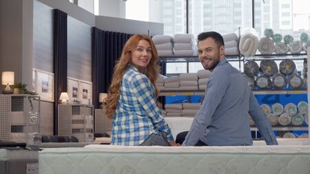 Cheerful couple smiling to the camera, while choosing new bed at furniture store. Beautiful woman and her husband sitting on orthopedic mattress at furnishings shop. Retail conceptの写真素材