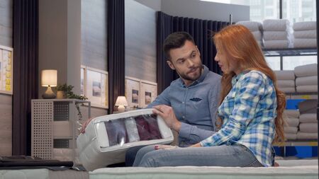 Beautiful couple choosing orthopedic mattress at furniture store together. Handsome man and his lovely wife discussing orthopedic mattress sample. Consumerism conceptの写真素材