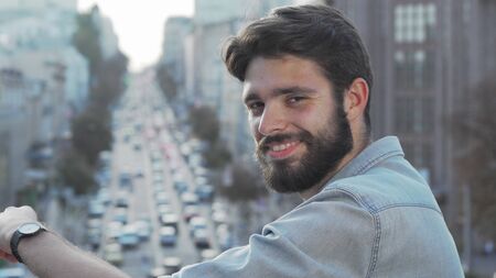 Handsome bearded man smiling to the camera over his shoulder. Young male hipster looking away at busy city streets, smiling joyfully to the camera. Success, achievement conceptの写真素材