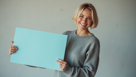 A joyful young woman with short blonde hair smiles brightly while holding a blank blue sign against a neutral background. Her warm expression and casual gray sweater create an inviting atmosphere, perfect for conveying messages or promotions.の素材