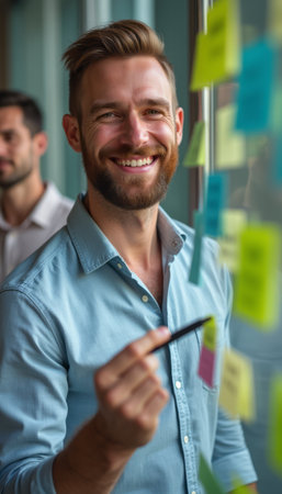 A cheerful man with a well-groomed beard is engaged in a brainstorming session, writing on colorful sticky notes. His bright smile radiates enthusiasm and creativity, capturing the collaborative spirit of teamwork in a modern office setting.の素材