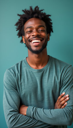 A cheerful young Black man stands confidently with his arms crossed, radiating positivity against a calming teal background. His bright smile and relaxed posture convey a sense of happiness and self-assurance, inviting viewers to share in his joy.の素材