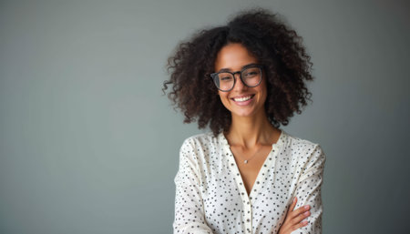 A joyful young woman with curly hair and glasses stands confidently against a soft gray background. Her bright smile radiates positivity and warmth, showcasing her stylish polka-dot blouse and vibrant personality.の素材