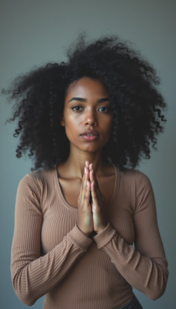 A young Black woman with voluminous curly hair stands in a prayer pose, exuding serenity and introspection. Her warm skin tone and focused expression create a powerful atmosphere of peace and mindfulness.の素材