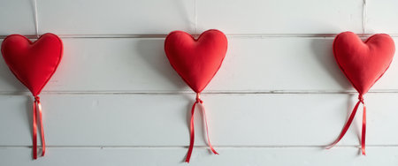 A trio of vibrant red heart-shaped cushions dangles elegantly against a clean white wooden backdrop, creating a striking contrast. The soft texture and delicate ribbons evoke feelings of love and warmth, making this image perfect for romantic themes and celebrations.の素材