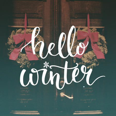 Hello winter text overlay on filtered photo with decor wreaths on the vintage door. Typography banner for greeting cards and social media content.の写真素材