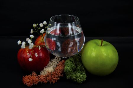 photo glass of water red and green apples moss different color white flowers on a black backgroundの写真素材
