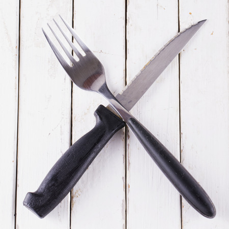 Black crossed cutlery over white wooden table, square image