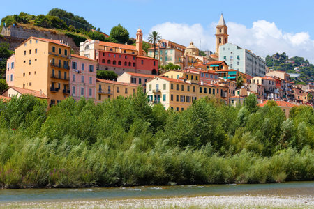 A view of a beautiful Italian old town Vintimille rising up into the hillside towards mountains in the distance. There are painted houses and church spire and wooded hills.の写真素材