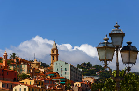 A view of a beautiful Italian old town Vintimille rising up into the hillside towards mountains in the distance. There are painted houses and church spire and wooded hills.の写真素材