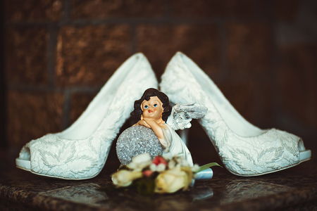 elegant white lace wedding bridal shoes high-heeled with a statuette on a dark brown background, morning of the bride.の写真素材