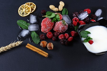 Frozen raspberry, strawberries, yoghurt plate mint leaves, blackberry, pieces of ice on shale board, slices of lemon, nuts, wheat, top view, close up setの写真素材