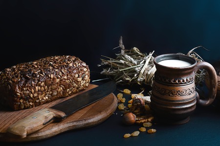 dark cereal bread with sunflower seeds, on a wooden board with a vintage knife, scalded nuts, cup of milk, raisins on shale board, concept of healthy eating, set.の写真素材
