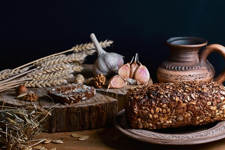 dark cereal bread with sunflower seeds, cup of milk, garlic and stems of wheat, nuts, concept of healthy eating, on a wooden background on a stump, set.の写真素材