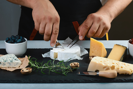 chef slicer camembert cheese with cheese knife on a black stone board with a twig of rosemary, walnut, Food banner for text or design. Side view photo, overhead. set.の写真素材