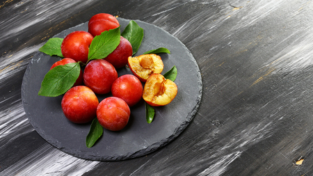 Red plum fruits on branch with green leaves on a round shale board a dark background. Top view, space for text.の写真素材