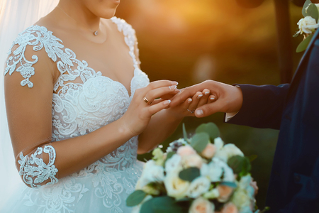 Beautiful wedding celebration at sunset. A bride in a white elegant dress holds the hand and puts the ring on her stylish bridegroom in a dark suit outdoors. Wedding wear, accessories.の写真素材