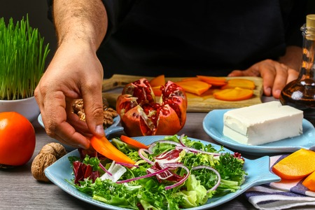 Close up of a chef hands preparing salad with persimmon, pomegranate, nuts and feta cheese. The concept of cooking recipe.の写真素材