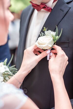 The bride's hand puts on a boutonniere flower on the groom's jacket. Bride puts a buttonhole on a grooms suit. close-up.の写真素材