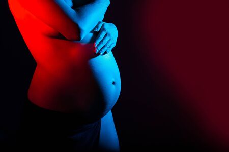 Closeup of belly of pregnant woman on gradient studio background in neon light. Concept of human...