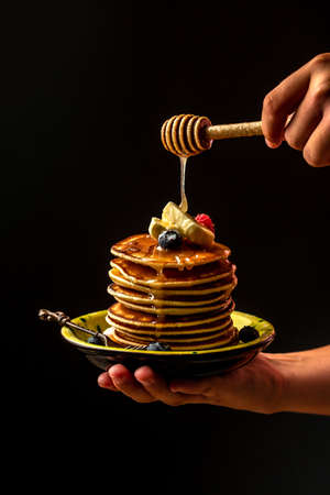 Woman pouring honey on tasty pancakes with berries on table, closeup. cooking by chef hand on dark...