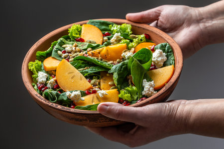 Woman holding vegetarian, detox bowl Fresh Tasty Persimmon salad with spinach, nuts, goat cheese, pomegranate, pumpkin seeds. Healthy balanced eating.の写真素材