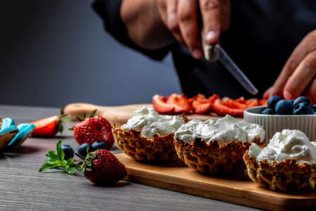 Hand with knife cuts strawberry. Berries on white cooking board. Fresh ingredients for a dessert. chef cooking berry dessert tarts with cream, strawberry and blueberryの写真素材