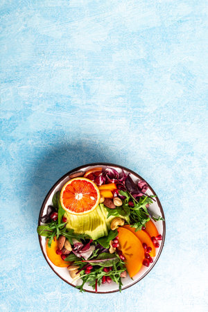 colorful buddha bowl with avocado, persimmon, blood orange, nuts, spinach, arugula and pomegranate on a blue background. vertical image, place for text.の写真素材