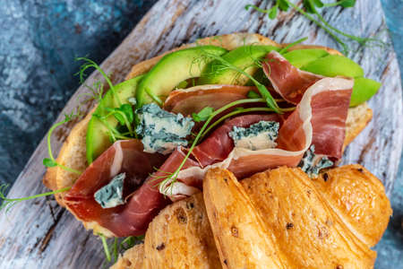 Croissant sandwich with prosciutto ham or jamon, blue cheese, avocado, microgin on blue background. Delicious snack.の写真素材