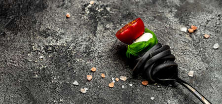 black pasta caprese salad tomatoes, mozzarella and green basil leaves on a black fork, Pasta with...