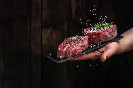 Chef salts steak in a freeze motion with rosemary and spices. Preparing fresh beef or pork on a dark...