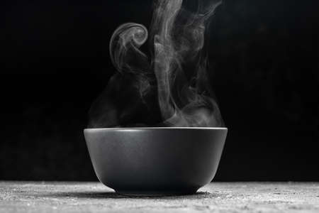 Bowl of hot steam of hot soup with smoke. black ceramic bowl on dark background. hot food. Culinary,...
