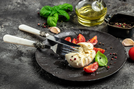 Salad with creamy Italian Burrata Cheese Served with Olive Oil, Basil Leaves and cherry tomatoes on white plate on a dark background. Restaurant menu, dieting, cookbook recipe top view.の写真素材
