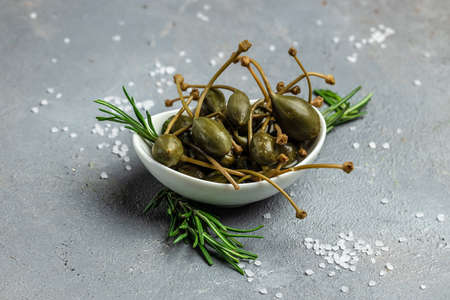 Capers. Marinated or pickled canned capers fruit with fresh rosemary in a small bowl, on gray background. place for text, top view.の写真素材