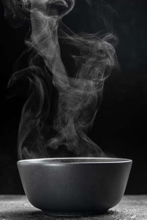 Bowl of hot steam of hot soup with smoke. black ceramic bowl on dark background. hot food. Culinary, cooking, concept.の写真素材