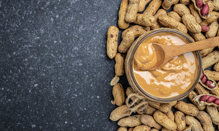 peanut butter or peanut paste in an open jar and peanuts in the peel scattered on a dark background,...
