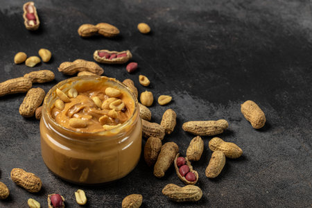 Peanut butter or paste in an open jar and peanuts in the peel scattered on a dark background,...
