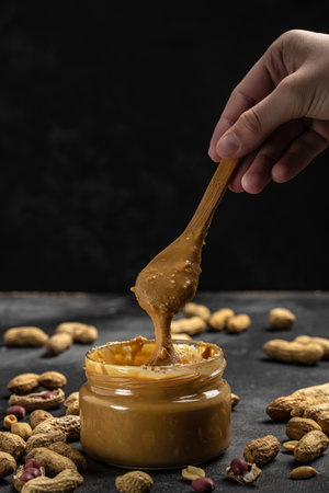 Peanut butter in glass jar and spoon on dark background, American dessert concept. vertical image....